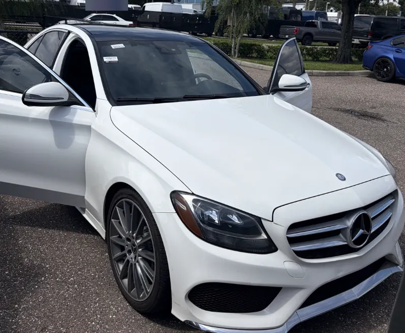 White Mercedes-Benz C-Class sedan with its door open in a parking lot during a windshield replacement